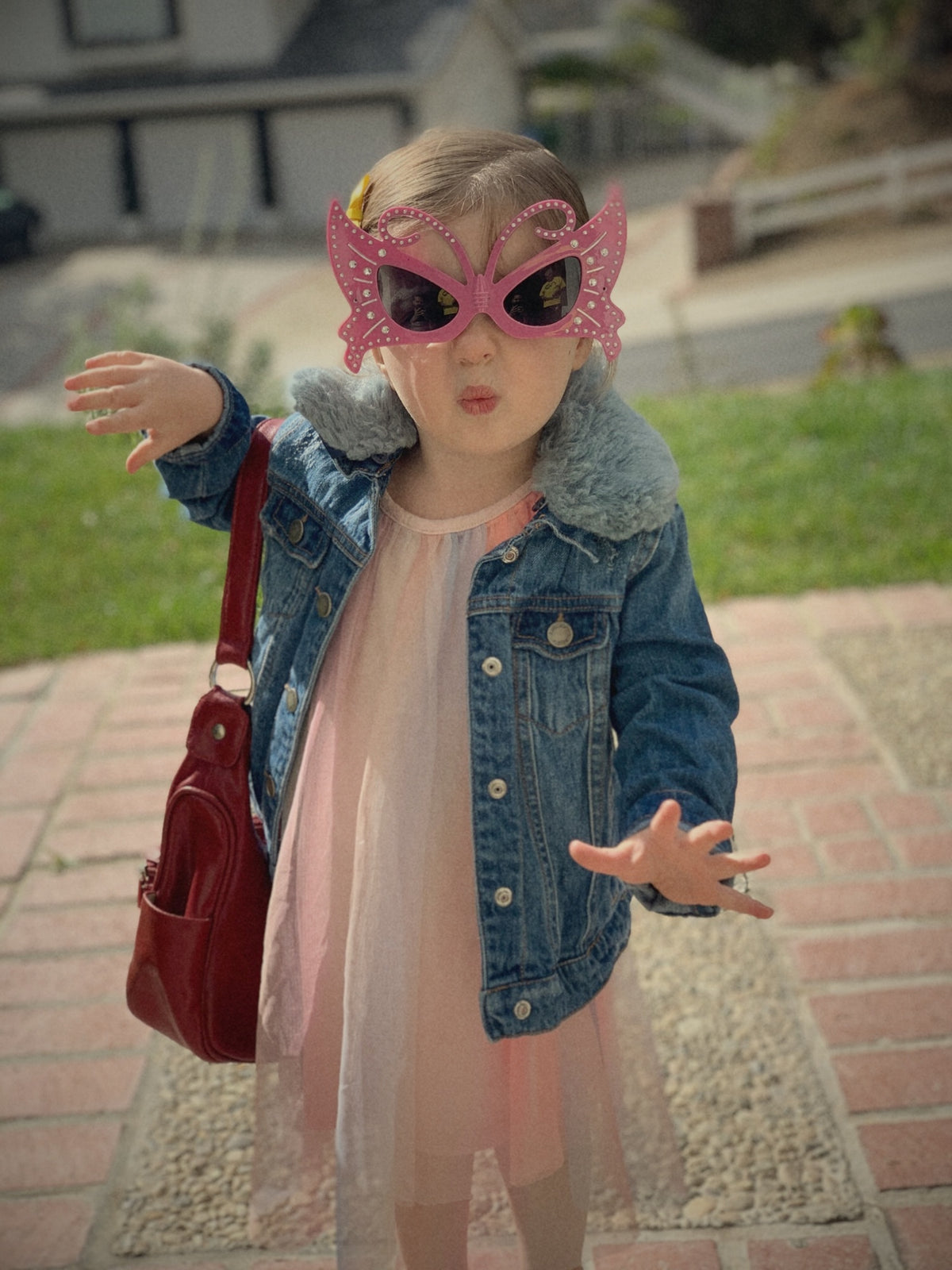 Afton's daughter as a toddler in pink butterfly sunglasses, a fur-collared denim jacket, a pink tulle dress and a red purse, blowing a kiss