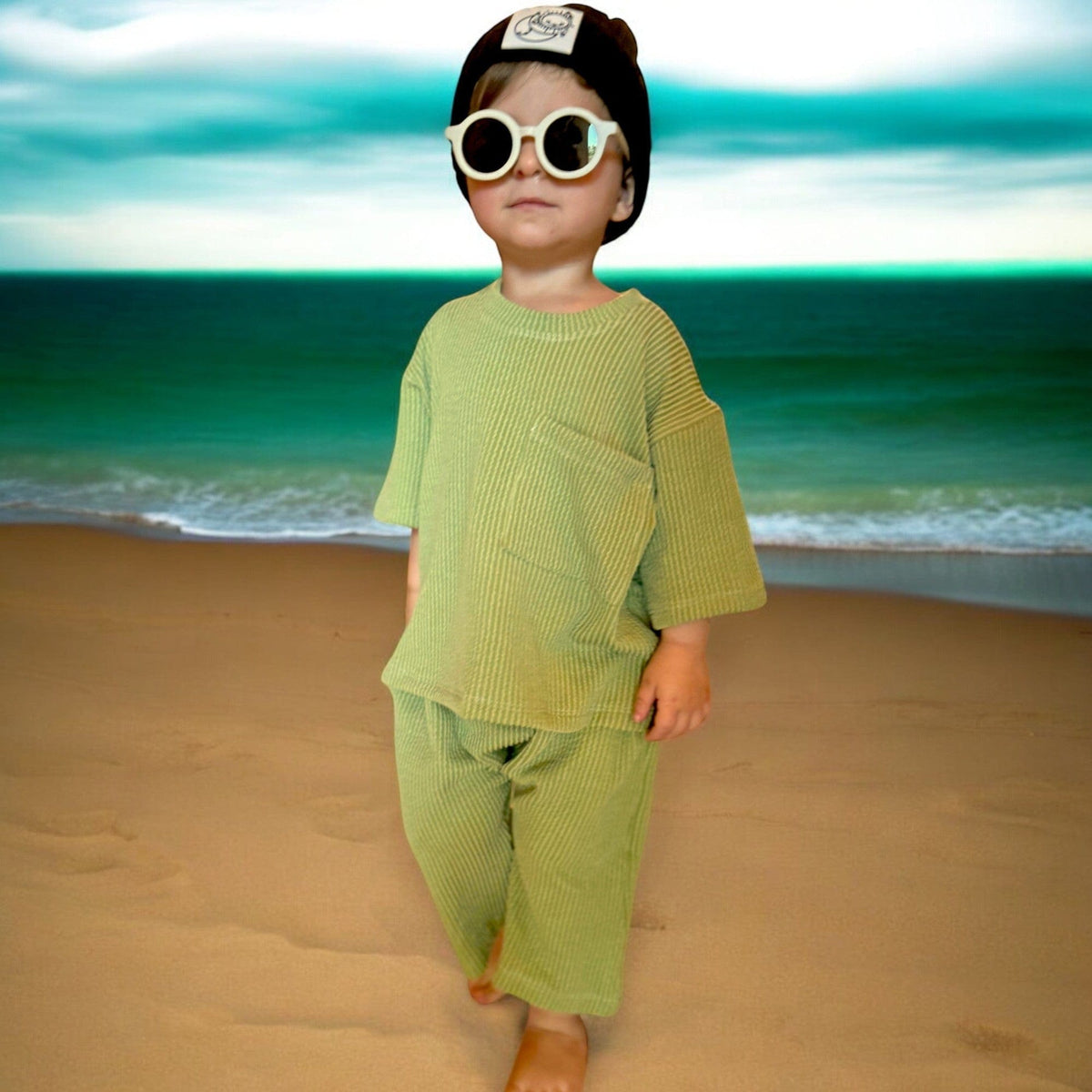 Toddler in white sunglasses on a beach wearing the matching Groovy Green tee and short