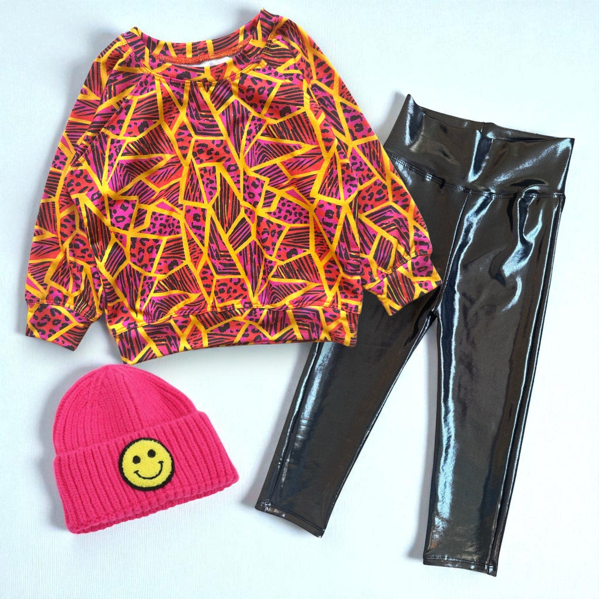 Neon Jungle Pullover laid flat with black metallic leggings and a pink smiley beanie