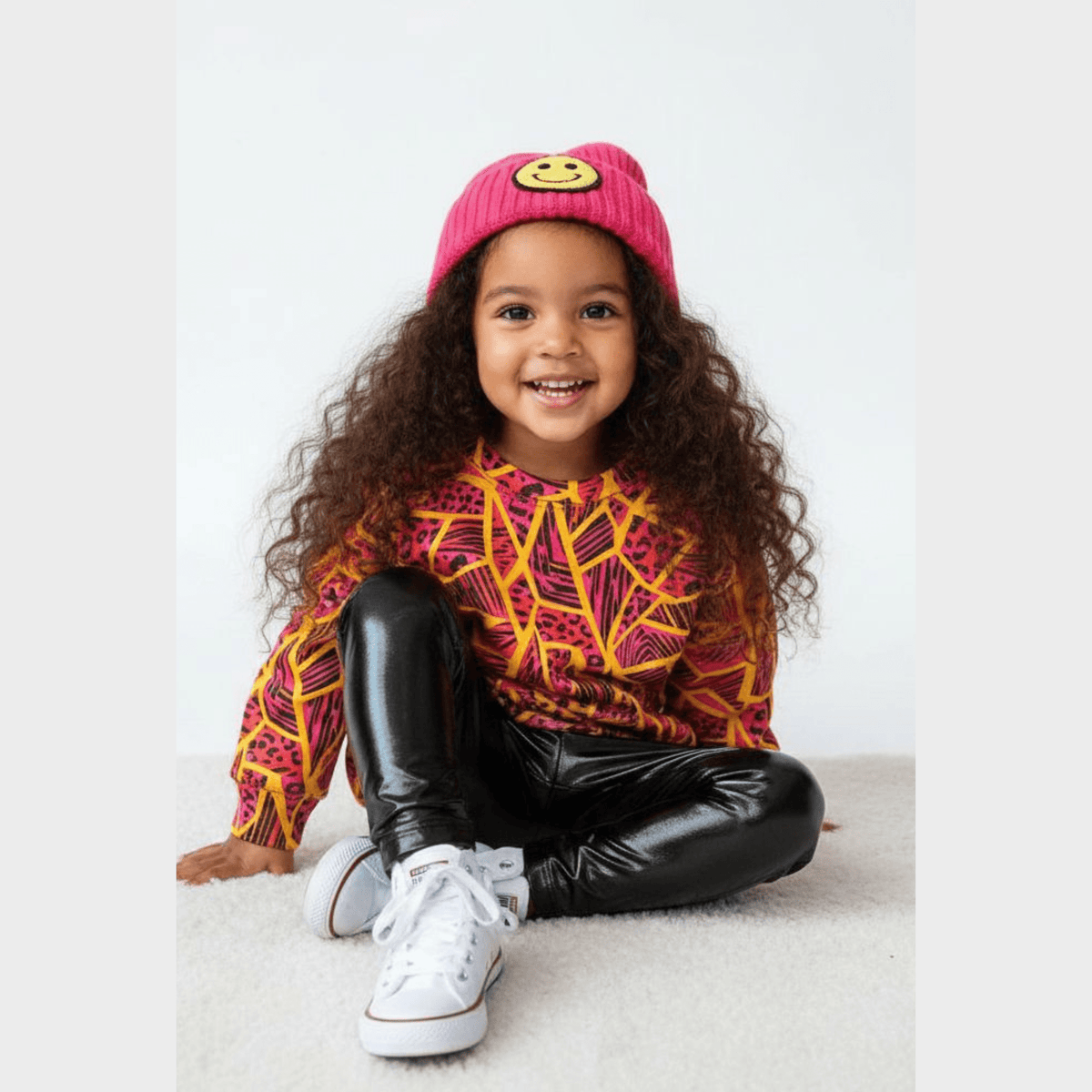 Smiling kid in a pink smiley beanie wearing the Neon Jungle Pullover and black metallic leggings