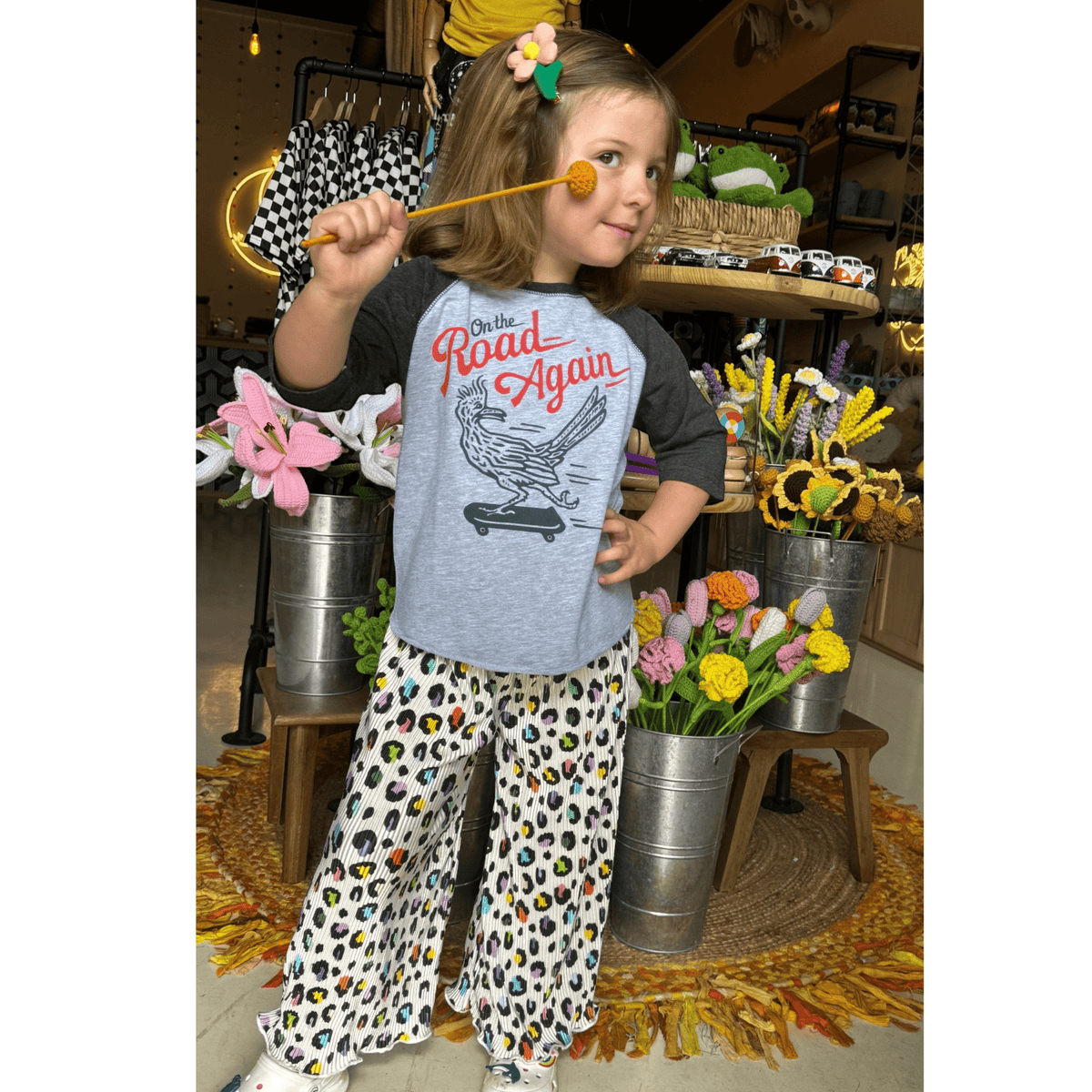 Kid in a graphic raglan tee and Loopy Leopard wide leg pants among buckets of flowers