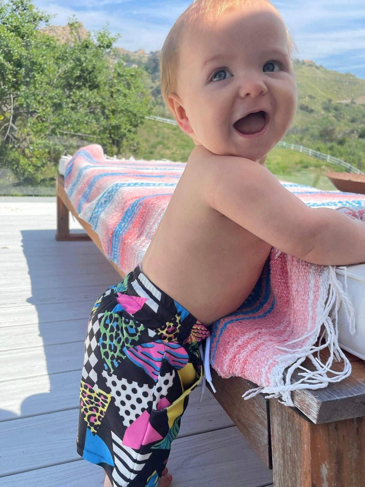 Smiling baby on a deck wearing Totally Rad swim trunks