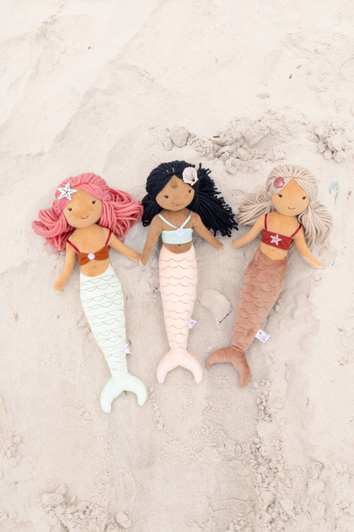 Three mermaid plush dolls with pink, black and blonde hair on the sand