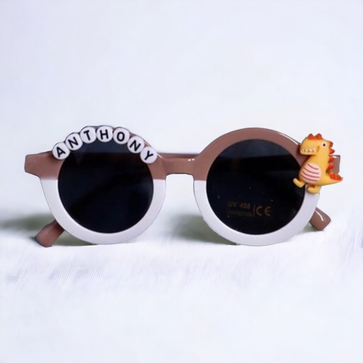 Brown and white sunglasses with a dinosaur charm and the name Anthony