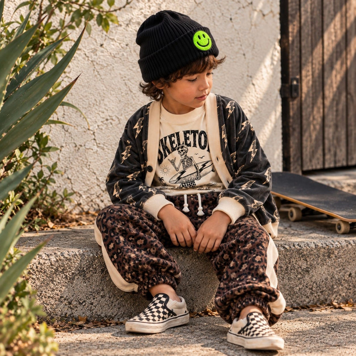 Boy sitting on a step in a smiley beanie, lightning-bolt cardigan, skeleton surf tee and leopard velvet sweatpants